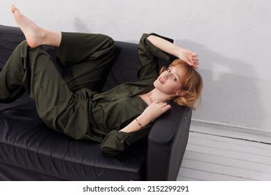 Portrait Of Young Girl In Dark Green Khaki Suit Laying On Black Sofa Barefoot. Pretty Blond European Girl With Short Hair-cut Smiling. Indoors. Doll Face Sexy Woman Relax At Home