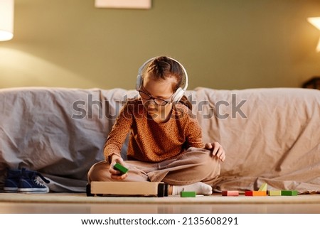 Portrait of young girl with down syndrome playing with toy blocks alone and wearing noise cancelling earphones, overstimulation concept, copy space