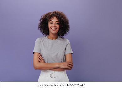 Portrait Of Young Girl In Gray T-shirt On Purple Background. Closeup Snapshot Of Curly Brunette Woman In Light Outfit Smiling On Isolated Backdrop