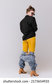 Portrait Of Young Girl Posing In Yellow Tights, Dropped Jeans And Boots Isolated Over Grey Studio Background. Weird Look. Concept Of Retro Fashion, Art Photography, Style, Queer, Beauty