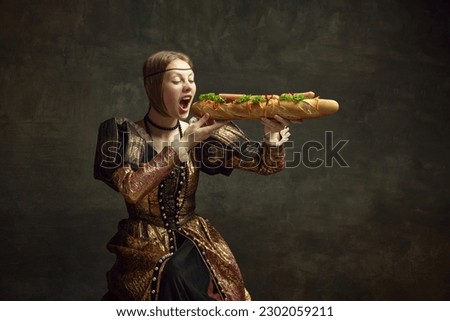 Portrait of young girl, princess in vintage costume eating giant baguette sandwich against dark green background. Concept of history, renaissance art, comparison of eras, health and food, diet