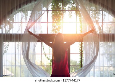 Portrait Of A Young Girl In A Red Dress And A Yellow Hat While She Is Opening A Curtain The Let The Light Come Through The Window.