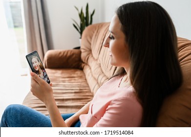 Portrait of young girl with a smartphone, she talks online with friend while sitting on the sofa at home. Concept of video communication on self-isolation, quarantine. Side view
