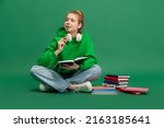 Portrait of young girl, student in casual cloth sitting on floor with thoughtful expression, studying isolated over green studio background. Concept of education, studying, homework, youth, lifestyle