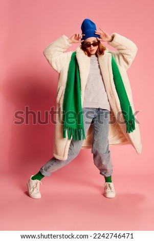 Portrait of young girl in sunglasses, blue hat, green scarf and fur coat posing over pink background. Stylish youth. Concept of beauty, winter fashion, lifestyle, emotions, facial expression. Ad