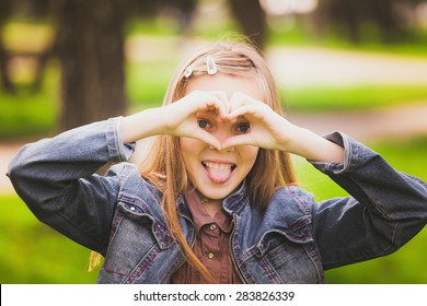 Portrait Of Young Girl With Tongue Out  Having Fun Outside. Funny Kid Looking Cheerfully At Camera Through Heart Made By Hands. Horizontal Color Photo.