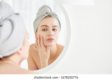 Portrait of  young girl with  towel on head in white bathroom looks and touches her face in the mirror and enjoys youth and hydration. Natural beauty, home care for problem skin