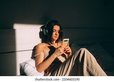 Portrait Of Young Girl Waking Up In Morning Listens To Music Using Headphones And Phone In Bed. Light Of Sun From Window Illuminates Girl In Bed.