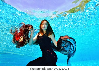 Portrait Of A Young Girl Who Swims And Plays Underwater In A Pool With A Red Cloth In Her Hands In A Red Dress On A Bright Sunny Day. Fashion Portrait. Underwater Photography. Horizontal Orientation.