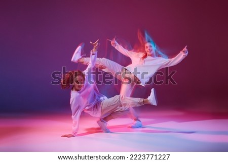 Portrait of young girls dancing hip-hop isolated over gradient violet background in neon with mixed light. Chaotic movements. Concept of movement, youth culture, active lifestyle, action, street dance