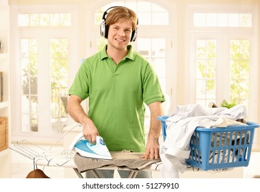 Portrait Of Young Goodlooking Man With Headphones Ironing Clothes In Living Room, Looking At Camera, Smiling.