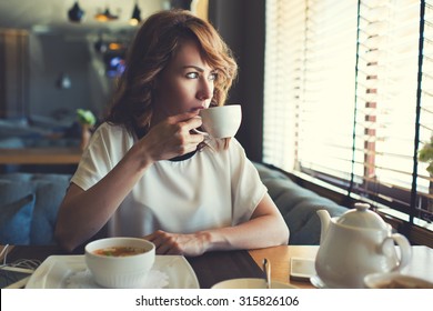 Portrait of young gorgeous female drinking tea and thoughtfully looking out of the coffee shop window while enjoying her leisure time alone, nice business woman lunch in cafe during her work break