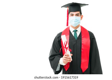 Portrait Of A Young Graduate Wearing A Face Mask And A Black Gown While Holding His College Certificate. Attractive Man Attending His Graduation During The Coronavirus Pandemic