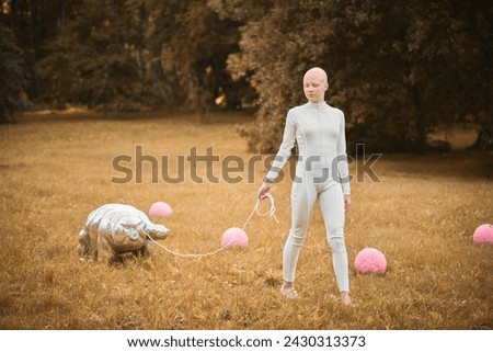 Portrait of young hairless girl with alopecia in white cloth walking tardigrade toy in fall park, surreal scene with bald teenage girl reflect on intertwining threads of life and art