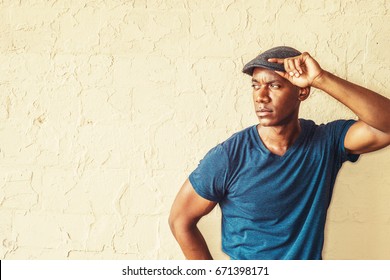 Portrait Of Young Handsome African American Man In New York. Wearing Blue V Neck T Shirt, Flat Cap, Black Guy Standing By Painted Wall On Street, Hand Touching Hat, Looking Away, Frowned, Thinking.
