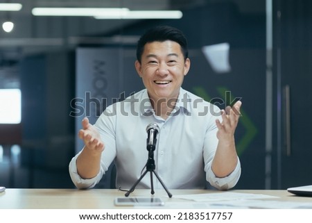 Portrait of a young handsome Asian man speaking to the camera into a microphone, recording a video message, conducting a webinar, making a podcast, gesturing with his hands, smiling in office
