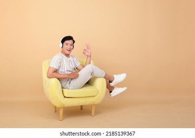 Portrait Of A Young Handsome Asian Office Man Sitting On Sofa Chair And Listening To Music Via Smart Phone, Concept Of New Normal Life Or Relaxing At Home As A Freelancer, Isolated On Background