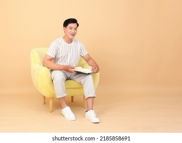 Portrait Of A Young Handsome Asian Office Man Sitting On Sofa Chair And Reading Book, Concept Of New Normal Life Or Work From Home As A Freelancer, Isolated On Background