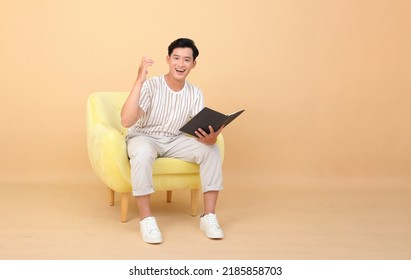 Portrait Of A Young Handsome Asian Office Man Sitting On Sofa Chair And Reading Book, Concept Of New Normal Life Or Work From Home As A Freelancer, Isolated On Background