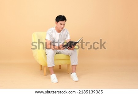 Portrait of a young handsome Asian office man sitting on sofa chair and reading book, concept of new normal life or work from home as a freelancer, isolated on background