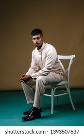 Portrait Of A Young, Handsome And Athletic Malaysian Indian Muslim Man In A Cream Colored Traditional Baju Melayu Tunic Sitting On A Chair. He Is Dressed Festively To Go Visiting For Raya. 