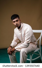 Portrait Of A Young, Handsome And Athletic Malaysian Indian Muslim Man In A Cream Colored Traditional Baju Melayu Tunic Sitting On A Chair. He Is Dressed Festively To Go Visiting For Raya. 