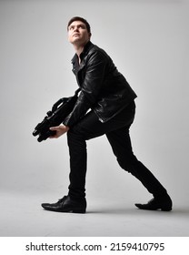 Portrait Of A Young Handsome Brunette Man Wearing Black Clothing And Leather Jacket, Holding A Scifi Gun Weapon. Dynamic Pose From A Low Angle Perspective Isolated On Studio Background.