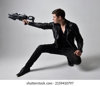 Portrait Of A Young Handsome Brunette Man Wearing Black Clothing And Leather Jacket, Holding A Scifi Gun Weapon. Dynamic Pose From A Low Angle Perspective Isolated On Studio Background.