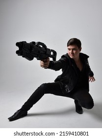 Portrait Of A Young Handsome Brunette Man Wearing Black Clothing And Leather Jacket, Holding A Scifi Gun Weapon. Dynamic Pose From A Low Angle Perspective Isolated On Studio Background.