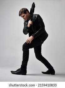 Portrait Of A Young Handsome Brunette Man Wearing Black Clothing And Leather Jacket, Holding A Scifi Gun Weapon. Dynamic Pose From A Low Angle Perspective Isolated On Studio Background.