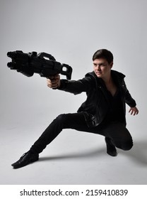 Portrait Of A Young Handsome Brunette Man Wearing Black Clothing And Leather Jacket, Holding A Scifi Gun Weapon. Dynamic Pose From A Low Angle Perspective Isolated On Studio Background.