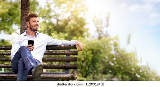 Portrait Of Young Handsome Business Man In Suit Relax On Bench In Park. He Holding Smart Phone And  Watches Thoughtfully In Green Forest Park. Escape In Nature To Recover Mind Business Concept.