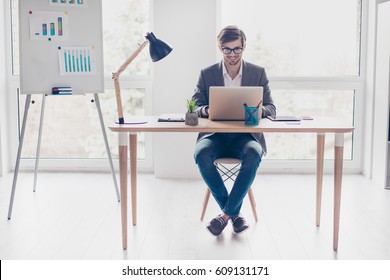 Portrait young handsome businessman with glasses is sitting at desk in workstation, smiling and typing on laptop.