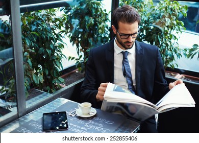 Portrait of a young handsome businessman reading a newspaper at his breakfast in coffee shop, brunette business man holding open newspaper sitting in cafe - Powered by Shutterstock