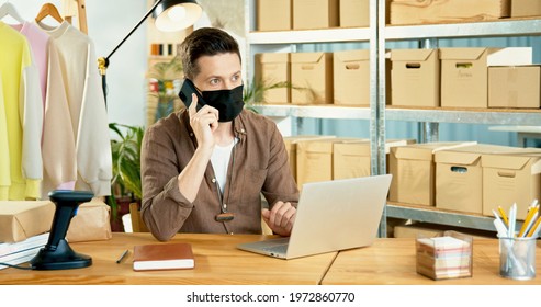 Portrait Of Young Handsome Caucasian Male Worker In Black Mask Sitting At Table And Calling On Smartphone While Browsing Online On Laptop. Man Seller At Clothing Store Working During Coronavirus