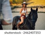 Portrait of young handsome cowgirl horseback riding at horse ranch and smiling at the cowboy in a blurry foreground. Smiling young attractive rider in western styled clothes is horseback riding.