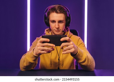 Portrait of young handsome cyber sportsman wearing headset playing online video game on his smartphone, looking at cellphone screen, focused on game, streaming. Playing mobile games, cyber sport - Powered by Shutterstock