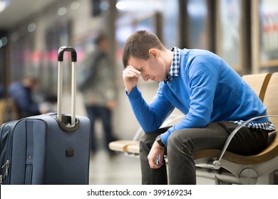 Portrait of young handsome guy wearing casual style clothes waiting for transport. Tired traveler man travelling with suitcase sitting with frustrated facial expression on a chair in modern station