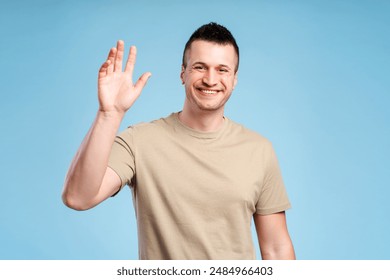 Portrait of young handsome man greeting or saying goodbye, waving his hand, standing isolated on blue background. Greeting concept