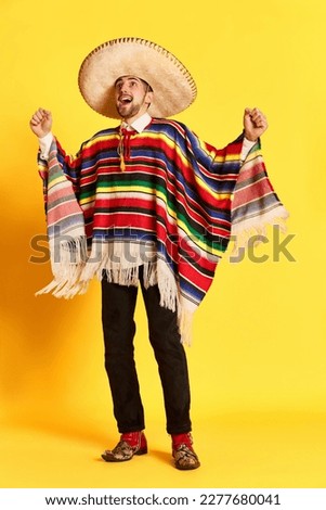 Portrait of young handsome man with happy excited face, in colorful festive clothes, poncho and sombrero posing against yellow background. Mexican traditions, celebration, festival, emotions concept