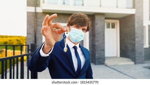 Portrait of young handsome man real-estate agent standing near new house and showing key. Male in medical mask holding house key. Selective focus on hands, close up.