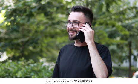 Portrait Of Young Handsome Man Talking On The Phone With Beard And Glasses. Smiling Young Man In Black Shirt. Outdoor Nature Background