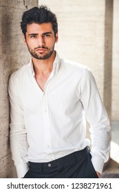 Portrait of young handsome man in white shirt outdoor. Nice appearance with stylish hair and beard. Leaning with a side on a wall