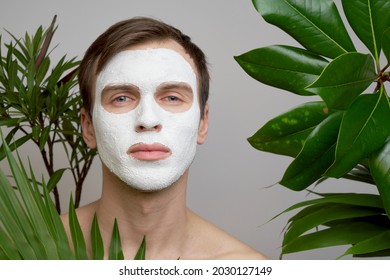 Portrait of young handsome man with white cosmetic mask on his face against background of green plants. Men's face care - Shutterstock ID 2030127149