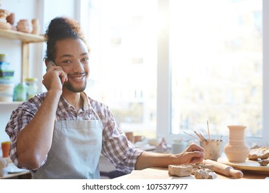 Portrait of young handsome mixed race male potter talking to customer on smartphone in workshop and smiling at camera - Shutterstock ID 1075377977