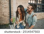 Portrait of young happy alternative urban couple in love holding bottles of cold fresh beverage and laughing on city street downtown. Smiling trendy hipster couple having fun time with refreshment.