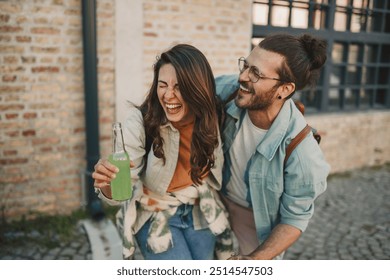 Portrait of young happy alternative urban couple in love holding bottles of cold fresh beverage and laughing on city street downtown. Smiling trendy hipster couple having fun time with refreshment. - Powered by Shutterstock