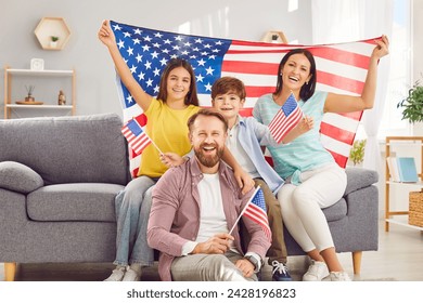 Portrait of a young happy american family with two kids sitting on sofa at home with flag of united states and looking cheerful camera celebrating Independence Day. Patriotic US holiday.