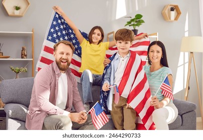 Portrait of a young happy American family with two kids sitting on sofa at home with flag of united states and looking cheerful camera celebrating Independence Day. Patriotic US holiday.