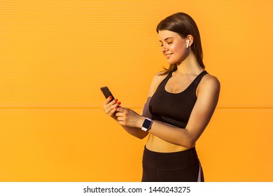 Portrait of young happy attractive sporty woman in black sportwear standing near orange wall background and holding phone, reading message with toothy smile, outdoor on summer, sunshine